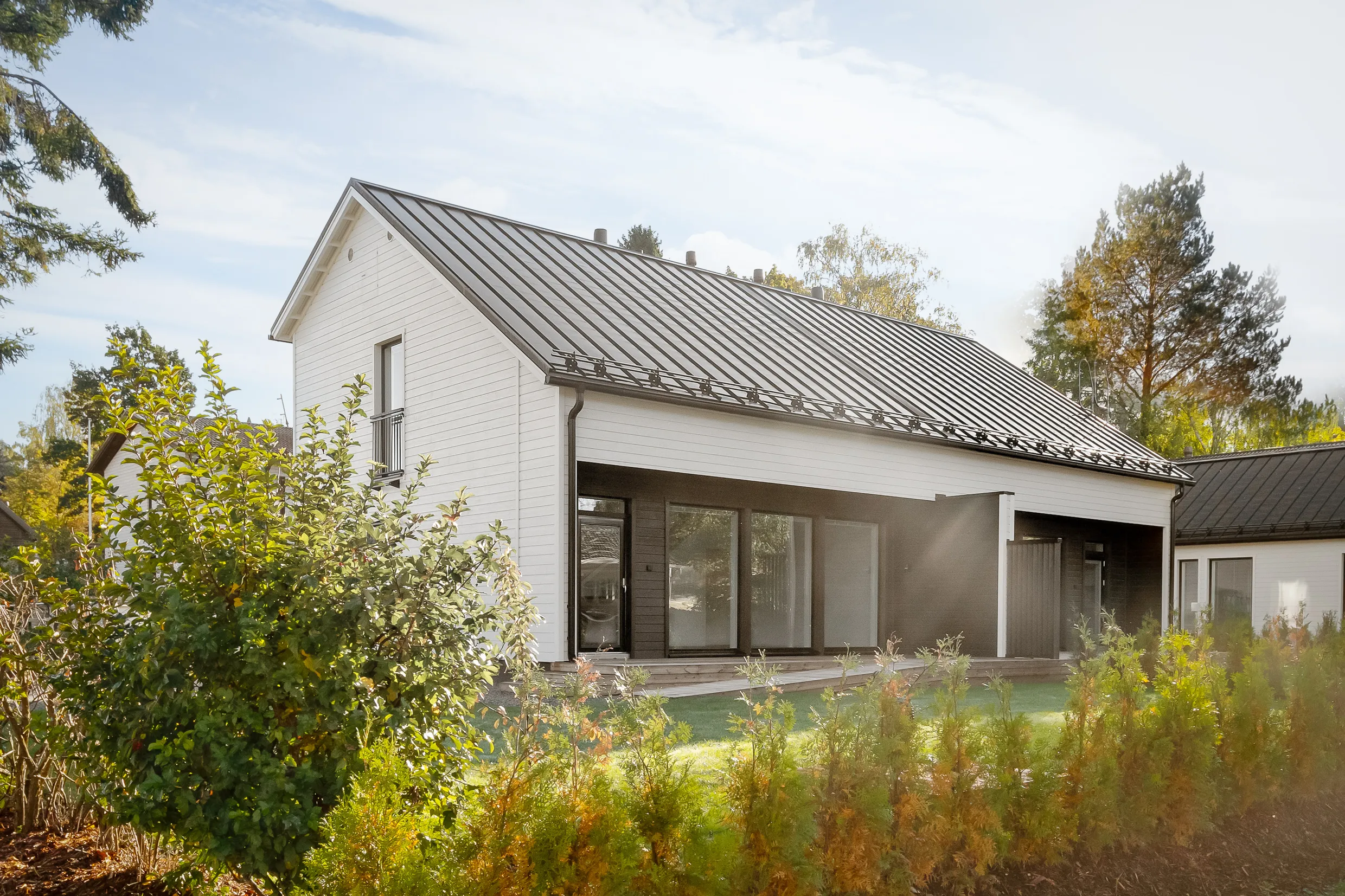 Moderni, valkoinen talo tummalla katolla, ympäröity vihreillä kasveilla ja puilla aurinkoisessa säässä.
